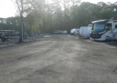 Multiple boats are stationed in a lot, organized in lines, with a backdrop of greenery and a clear sky.